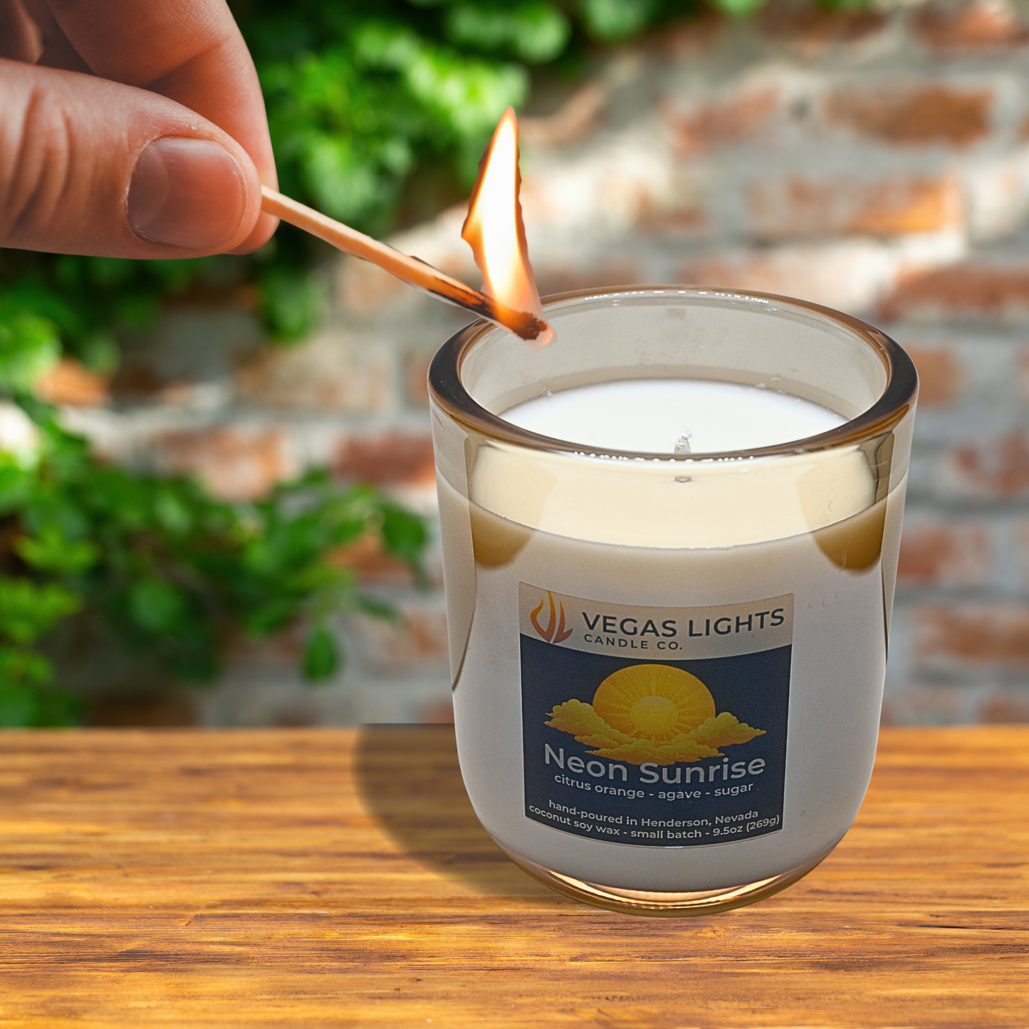 Person lighting a candle labeled 'Vegas Lights Candle Co. Neon Sunrise' with a blurred natural background.